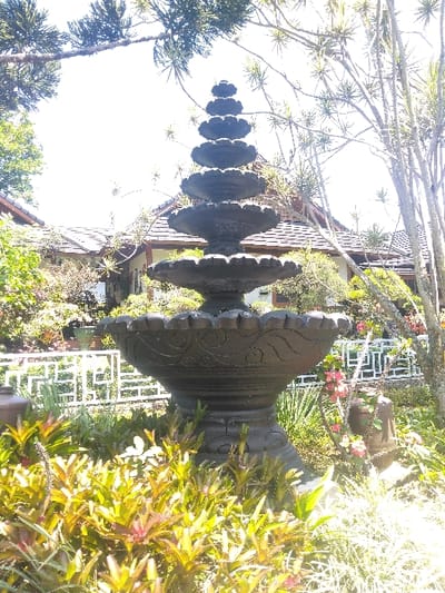 A dark, multi-tiered ornamental fountain, featuring seven visible scalloped basins decreasing in size towards the apex, stands centrally in a brightly lit outdoor garden scene. The large bottom basin is adorned with sculpted swirl patterns. Lush green and reddish-green foliage occupies the immediate foreground, surrounding the fountain's base. Behind the fountain, a white metal fence with geometric patterns is visible, followed by several buildings with light-colored walls and dark, sloped, shingled roofs. Various trees and shrubs are scattered throughout the background, with the bright, overexposed sky visible above. No individuals are present. The scene is located in Nová Bystřice, Czechia.