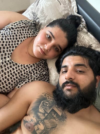 This is a close-up, overhead shot of a couple lying in bed, likely in Mainburg, Germany. The woman, on the left, is lying on her back with her head resting on a patterned pillow. She is wearing a black and white patterned top. Her hair is pulled back in a bun. The man is lying next to her, on his right side, facing the camera. He has a full beard and a tattoo of a lion and praying hands on his chest. He is shirtless. The background consists of a pillow and what appears to be the edge of a bed. The lighting is soft and natural.