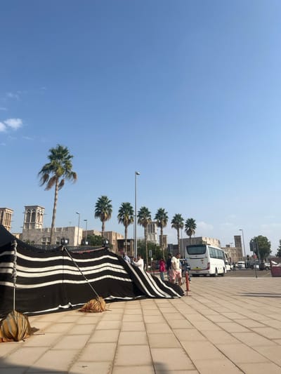 The scene depicts an outdoor urban environment within Deira, United Arab Emirates, under a clear blue sky with minimal cloud cover. In the foreground, a large, low-lying tent or canopy structure is visible, constructed from black fabric with prominent horizontal white stripes. This structure is secured to the light-colored paved ground by two distinct, decorative, possibly weighted objects, one on the left and one centrally located.
In the midground, several individuals are present. Multiple people, including men and women in casual attire (e.g., shorts, t-shirts), are standing near the tent's edge and further back, observing the surroundings. A white passenger bus (coach) is positioned on a paved roadway to the right, facing away from the viewer. A tall, light-grey street light pole stands prominently in the center-right.
The background features a row of mature palm trees with green fronds and visible trunks, lining a street. Behind the palm trees and further back are several traditional-style buildings constructed from light-colored materials, characterized by distinct square wind towers (Barjeel) on their rooftops. Additional structures, vehicles, and street infrastructure are visible in the far background. The overall impression is a sunny day in an area blending historical architecture with modern urban elements.