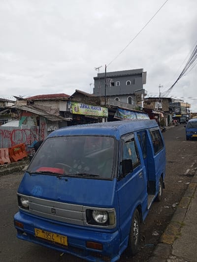 A blue Suzuki Carry minibus sits parked on a narrow street next to a curb. The vehicle's license plate reads "F 1954 YT". Behind it, a cluttered street scene unfolds with aged buildings, utility poles, and wires crisscrossing the sky. A sign reading "JAWA MANUK" is visible among the buildings. In the distance, another similar blue vehicle can be seen. The sky is overcast.