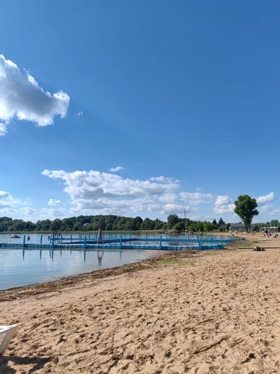 The image depicts a sunny day at a beach on a lake in Myadzyel, Belarus. The foreground is dominated by a sandy beach with footprints visible. In the middle ground, a bright blue floating platform extends into the calm lake, with several people walking on it and in the water. A red inflatable raft is also visible on the lake in the distance. The background features a dense line of green trees and some buildings. The sky is a clear blue with scattered white clouds. The overall scene conveys a sense of leisure and recreation.
