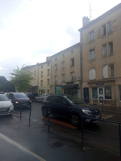 The image shows a street in Nancy, France, with several cars stopped in traffic on a wet road. The buildings on the right are lined with windows and shutters, and one building has an "Allianz" sign above its entrance. A tree is visible on the left side of the image, and the sky is overcast. The cars are facing forward, indicating they are moving in the same direction. The road appears to be a city street with sidewalks and streetlights.