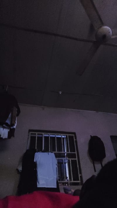 The image depicts an interior scene, likely a bedroom, at night or in low light conditions. A ceiling fan is visible at the top of the frame. Below the fan, on a wall, a barred window is present, with what appears to be clothing hanging in front of it and to the left. A white t-shirt is prominently displayed hanging on a rack. To the right of the window, a black backpack is hanging on the wall. The foreground is occupied by a red blanket or fabric. The scene is set in New Nyanya, Nigeria.
