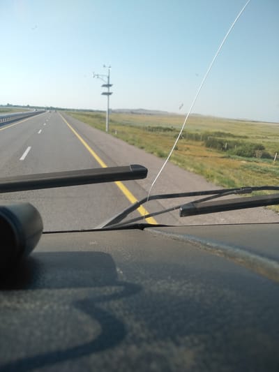 A low-angle shot, taken from inside a vehicle, looks out through the windshield at a highway stretching into the distance under a clear, pale blue sky. The highway, with a yellow line dividing lanes and white dashed lines marking lanes, is bordered by dry, grassy fields and a few scattered bushes. A tall, utility pole with various instruments is visible on the right side of the road. The foreground is dominated by the textured dashboard of the vehicle and the dark arm of a windshield wiper. A prominent crack runs diagonally across the windshield. The scene appears to be in a rural or steppe environment, consistent with the location of Баймырза, Kazakhstan, known for its open landscapes and agricultural areas.