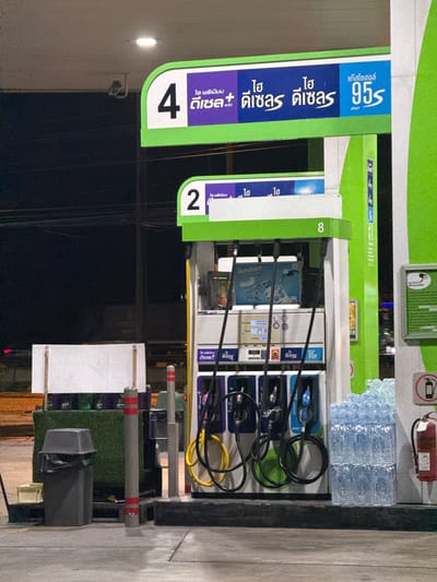 The image shows a gas station at night in Sanap Thuep Subdistrict, Thailand. The gas station features multiple fuel pumps with digital displays and hoses. Signs above the pumps indicate fuel types and prices in Thai script. To the left of the pumps, a grey trash can is visible. To the right, a stack of bottled water and a fire extinguisher are placed near the pumps. The ground is paved with concrete. The overall lighting suggests it is nighttime, with artificial lights illuminating the scene.
