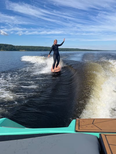 An adult female is wake surfing on a body of water. She is wearing a black wetsuit and is standing on a light brown surfboard. She is positioned in the center of the frame, riding the wake created by an unseen boat. The water is dark and choppy, with white foam from the wake. In the background, a dense green forest lines the opposite shore, and a bright blue sky with wispy white clouds stretches overhead. The lower right portion of the image shows a part of a boat's deck and railing. The scene takes place outdoors on a sunny day.