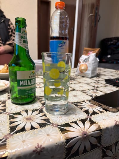 The image shows a tabletop scene. A green glass bottle labeled "Младо Пирински" is lying horizontally on the patterned tablecloth. Beside it, there is a transparent glass with a lemon design. An orange-capped bottle of what appears to be "Nestea" is slightly behind the glass, along with a white package of chips, which is placed further back. A cellphone lays on the table. In the background, there's a glimpse of a doorway and what appears to be a backpack. The details on the green glass bottle indicate it is produced by "Carlsberg Bulgaria" and the best before date is on the bottle. The text on the bottle also reveals that it's a "Непастьоризовано" (unpasteurized) drink. While the photo does not explicitly state its location within the city of Popintsi, Bulgaria, the fact the brand is a product of Carlsberg Bulgaria gives a hint to the country of origin and therefore might be taken in any location in the country of Bulgaria.