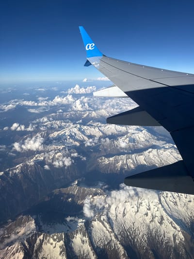 The image shows an airplane wing with a blue and white logo flying over a mountainous region. The wing is partially illuminated by sunlight, with the rest in shadow. Below the wing, there are snow-capped mountain peaks with varying shades of brown and gray. The sky above the wing is a clear blue. This scene is not identifiable with any specific location in the city of Mayrhofen, Austria, due to the view being from an airplane above the terrain. The environment shows a cold, mountainous region, likely a part of the Alps. There are no people visible in the image.