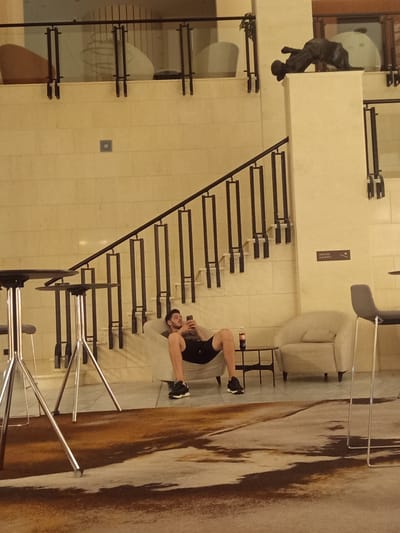 A man is sitting in a chair holding a phone in his left hand and looking at it. He is wearing black shorts and black and white sneakers. A bottle of Coca-Cola is on a small table to his right. The man is in a room with a staircase and a chandelier. The location is the city of Razliv, Bulgaria, in a hotel lobby.