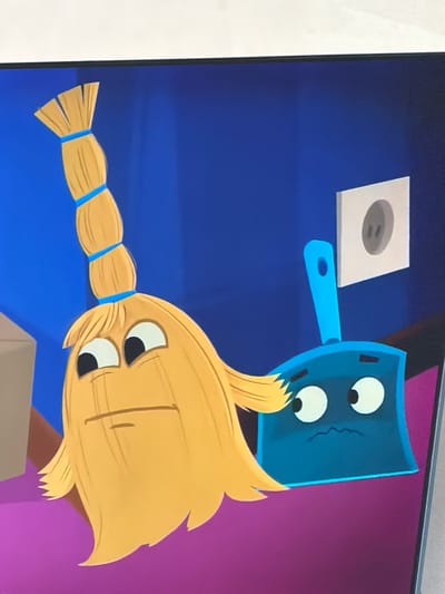The image is a close-up of an animated scene featuring two anthropomorphic cleaning tools against a blue background. On the left is a yellow broom with a segmented handle tied with blue bands and a head of bristly yellow strands that form a face with wide, surprised eyes and a straight mouth. Next to it on the right is a blue dustpan with a similar cartoon face, featuring large, round white eyes with black pupils and a small, wavy, distressed mouth. A light gray electrical outlet with two dark gray holes is mounted on the blue wall to the right of the broom. The lower portion of the scene shows a purple surface, and to the far left, a corner of a brown cardboard box is visible. There is no visible connection to the city of Krasnyi Krym, Russia.