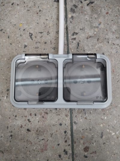 An outdoor, double electrical socket is mounted on a speckled concrete surface. A white conduit extends vertically upwards from the top of the socket. Each socket is protected by a clear, hinged plastic cover. The socket housings are light grey. The right-hand socket has "IP 54" printed on its cover. The concrete appears to be part of a sidewalk or paved area, with a visible seam running vertically down the center.