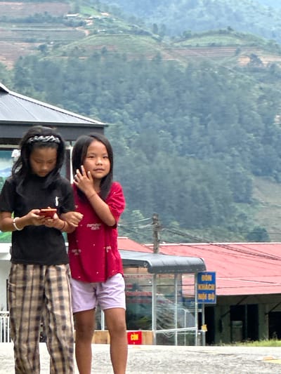 This image depicts a scene in Pung Luong, Vietnam, featuring two young Asian girls standing outdoors with a mountainous landscape in the background.

The girl on the left, with long dark hair, wears a grey and white patterned headband, a black short-sleeved t-shirt with a small white graphic on the left chest, and light-colored plaid pants. A light-colored bracelet is visible on her left wrist. She is looking down, focused on a red smartphone held in both hands. The girl on the right, also with long dark hair, wears a red short-sleeved t-shirt with subtle white text (possibly "SUMMER") and light-colored shorts. A light-colored bracelet is on her right wrist, and her right hand is raised near her face. She is looking directly towards the camera. The girls appear to be linking arms or holding hands, indicating interaction.

In the mid-ground behind the girls, a building with a dark corrugated roof is visible. Further back, several structures with red corrugated roofs are present. A blue sign with white Vietnamese text reads "ĐÓN KHÁCH NỘI BỘ" (Welcome Local Guests) and is attached to a metallic structure, possibly a shelter. A red sign with partially visible white text ("CON" or "CÔN") is on the ground nearby. A wooden utility pole with multiple dark wires stands to the right. The background features multiple layers of green, heavily forested mountains under a hazy sky, with some cleared or terraced sections on the slopes. The girls are standing on a paved or concrete surface.