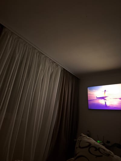 The image shows a dimly lit room with a large television screen displaying a sunset scene with a person on a boat. To the left, floor-to-ceiling curtains are drawn, partially obscuring the wall. The television is mounted on the wall and is the primary source of light in the room. In the foreground, a patterned pillow is visible, suggesting a seating area. The environment appears to be a residential space. No specific location in Novosilka, Ukraine, is identifiable from the visual information provided in the image.