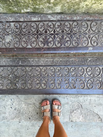 This media file features a close-up overhead shot of a person's feet wearing tan Birkenstock sandals, positioned on a concrete surface. The feet are adorned with golden-brown nail polish. To the left of the feet, there is a textured, ornate metal grate with intricate scrollwork and floral motifs. The grate appears weathered and set against a rough, moss-covered stone wall on the far left. The scene suggests an outdoor setting, likely a step or walkway with architectural details. The lighting indicates it is daytime, with no specific weather conditions discernible. The overall impression is one of an urban or historical environment, possibly in Taganrog, Russia, given the location context. The focus is on the juxtaposition of the modern footwear against the ancient-looking metalwork and rough concrete. There is no visible text in the image.