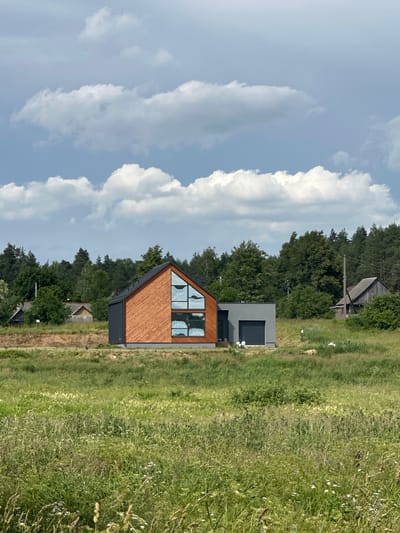 The image depicts a modern, triangular-shaped house with wooden siding and large, multi-paned windows on its front facade. The house is situated in a rural setting, with a field of tall, green grass and wildflowers to its right and a dense line of trees in the background. To the left of the house, the sky is filled with dramatic, puffy white clouds against a pale blue backdrop. A smaller, more rustic wooden structure is visible further back among the trees, suggesting the presence of other buildings in the area. The scene appears to be set during the daytime, with bright, natural light casting subtle shadows, indicating fair weather. The overall mood is tranquil and serene, highlighting the modern architecture within a natural landscape. No text is visible in the image.