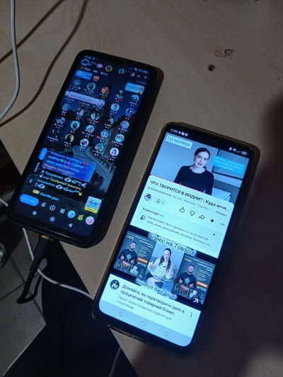 Two smartphones are placed on a light brown wooden surface. The phone on the left is black and shows a live streaming interface with numerous small circular profile pictures of users, along with text in Russian, indicating a chat or audience feed. A notification bubble in Russian announces a reward for watching for 60 seconds. The phone on the right is also black and displays a YouTube video interface. The video features a woman speaking, with text overlays in Russian, and below it, a title that translates to "What is happening in India? Where else..." and a comment section. Further down, another video preview with the title "Business on Goods" is visible. The scene is dimly lit, and a white cable is present on the left side of the image, partially obscured by the left phone. The background is out of focus. The location in Chutove, Ukraine is not ascertainable from the image.