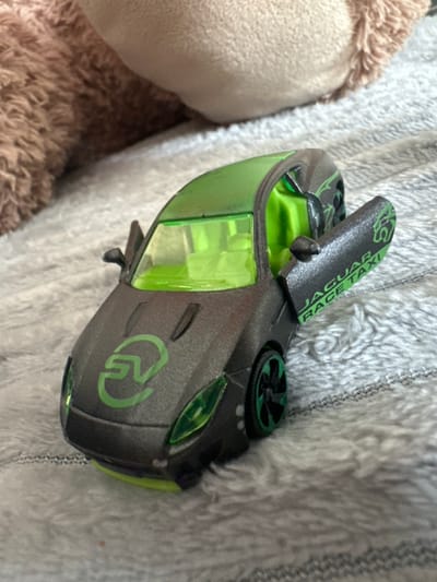 An indoor scene displays a dark grey toy car with neon green accents, positioned on a light grey, textured, fuzzy blanket. The vehicle features transparent neon green windows, an illuminated green interior, and green wheel rims. A stylized green 'S' or 'G' logo is present on the front hood. The driver's side door is open, revealing the words "JAGUAR RACE TAXI" printed vertically in green on its inner surface. A large, out-of-focus, brown, plush object occupies the upper portion of the background. No specific geographical markers identifying a precise location within Easingwold, United Kingdom, are discernible in the image.