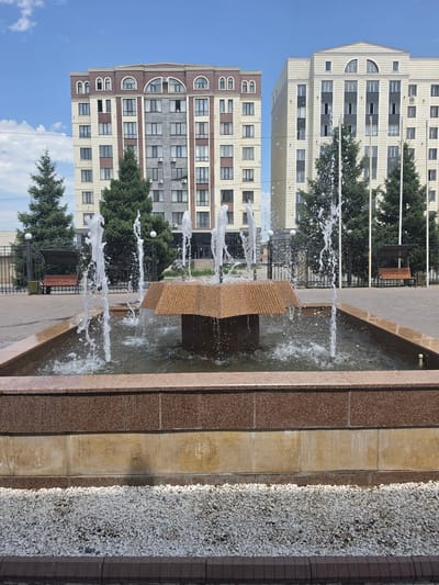 This is a vertical, eye-level shot of an outdoor fountain with multiple water jets. The fountain is constructed with granite and stone, and surrounded by gravel and brick. In the background, there are several multi-story buildings, some with arched windows. Green trees are visible between the fountain and the buildings. The sky is blue with some clouds. The scene appears to be a public space, possibly a park or plaza, in Karakol City, Kyrgyzstan. The lighting suggests it is daytime, likely late morning or early afternoon. There is no visible text in the image.