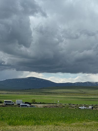 The image captures a landscape in or near Tlik, Armenia. Dominating the left of the frame are dark, ominous storm clouds. Adjacent to the clouds is a mountain range with patches of snow on its peaks. To the right of the mountains is a large expanse of green, grassy land. A road runs parallel to the mountain range, along which several white vehicles are parked. The setting conveys a sense of rural or semi-rural location and a dramatic weather event.