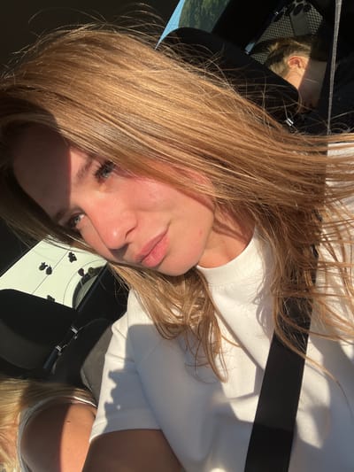 A blonde-haired woman is in a car, wearing a white shirt and a seatbelt. The location is implied to be Palje Brdo, Croatia.