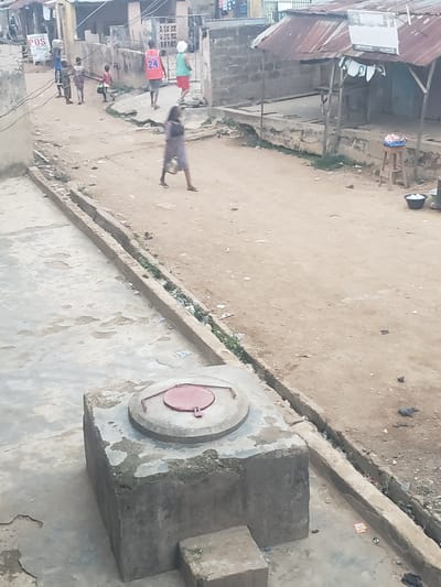 The image shows a street scene in Alalubosa, Nigeria, viewed from an elevated position. Several individuals are present on the street. One person appears to be walking alone, while others are clustered together. Some individuals are wearing brightly colored shirts. There is a concrete drainage ditch running along the edge of the street. A large, square concrete structure with a circular, red-lidded opening sits near the foreground. The surrounding buildings are made of concrete and corrugated metal. The environment seems dusty.