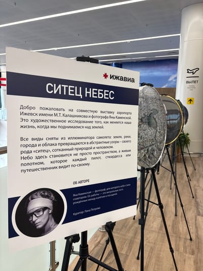 This media file features a sign with Russian text displayed at what appears to be an airport or exhibition space. The sign has a dark blue and white design and prominently displays the title "СИТЕЦ НЕБЕС" (Sitets Nebes), which translates to "Cotton Sky." The text describes a joint exhibition by the Izhevsk Airport named after M.T. Kalashnikov and photographer Yana Kamenskaya. It explains that the exhibition showcases how life changes as we ascend above the earth, with aerial views of landscapes transforming into abstract patterns, like a "cotton sky" woven by nature and people. The sign also includes information about the author, Yana Kamenskaya, as a photographer who collaborates with the sky, and the curator, Lana Petrova. Below the text, a black and white circular image is visible, depicting the profile of a person with their eyes bandaged, wearing glasses. The background of the image shows various black tripod stands, suggesting an exhibition setting. To the left of the sign, a vertical white panel with the word "ВЫЛЕТ" (Departure) and an airplane icon is visible, further reinforcing the airport context. The lighting in the scene appears to be artificial, possibly from overhead fluorescent lights.