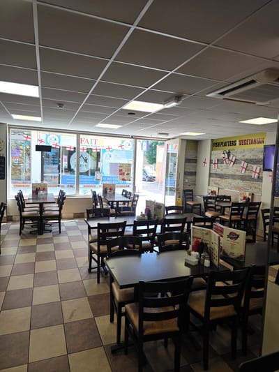 This is an interior shot of a cafe in King's Lynn and West Norfolk, United Kingdom. The cafe appears to be a fish and chip shop or similar establishment. The space is furnished with dark wood tables and chairs, with some tables already set with condiments. The floor is tiled with a checkered pattern of brown and beige squares. The walls are decorated with a stone-like tile and several English flags are hung on the wall and in the window. The window displays advertising for "FAITH Community Healthcare Ltd." and mentions opening hours. A sign above the counter reads "FISH PLATTERS VEGETARIAN". The overall atmosphere is bright and well-lit, with fluorescent lighting and natural light coming from the windows. There are no people visible in the image. The image captures the interior of a business that appears to be open for customers.