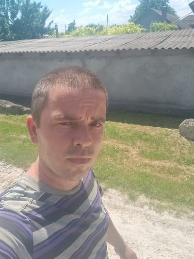 A medium shot captures a man in an outdoor setting, his face turned towards the camera. He is wearing a t-shirt with horizontal stripes in shades of purple and gray. The background features a mix of green grass, a gray, wavy roof, and trees under a bright blue sky, suggesting a daytime scene with clear weather. A rough, light-colored surface, possibly gravel or dirt, is visible in the foreground. The man's expression is neutral. A closer inspection of his t-shirt reveals some faint, possibly printed text on the shoulder area. The overall impression is that of a casual, outdoor moment. The location context provided is Mitoc, Moldova.