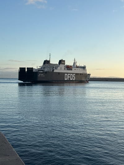 This media file shows a large ferry ship named "KATTEGAT" sailing on the water. The ship has "DFDS" prominently displayed on its side. The ship is dark gray with white structures on top and life preservers visible. The water is choppy, reflecting the sky. In the background, there is a faint outline of land with a line of trees or structures along the coast, and a hazy sky that suggests either early morning or late afternoon. The overall scene is peaceful and conveys a sense of maritime travel. Visible text includes "DFDS", "KATTEGAT", and "GIBRALTAR". The orientation of the image is such that the ship appears to be sailing vertically.