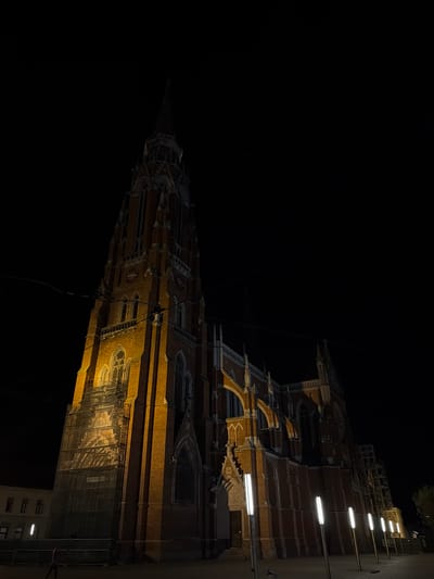 A nighttime view, rotated to the right, features the illuminated spire of the Co-cathedral of St. Peter and St. Paul in Osijek, Croatia. The structure is lit with warm-toned lighting, showcasing its architectural details. Scaffolding is visible near the base of the spire, suggesting ongoing maintenance or restoration. Some lines of electric light fixtures, running vertically along an adjacent building, are clearly visible. The sky is mostly black, indicating a dark, nighttime setting.