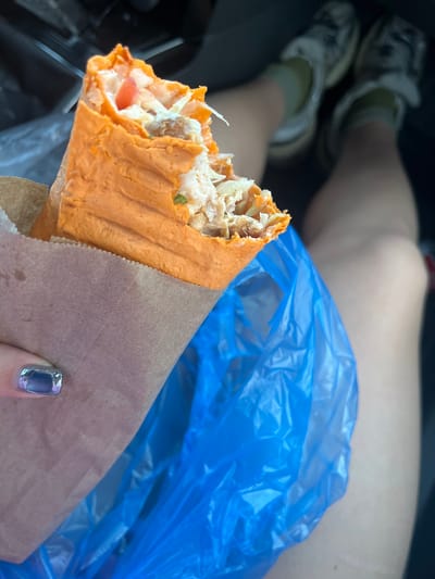 The image captures a close-up of a person's hand holding a shawarma, wrapped in brown paper, with a bright blue plastic bag in the background. The shawarma appears to be made with a reddish-orange flatbread, filled with shredded meat, white sauce, and some visible pieces of tomato and greens. A person's legs, wearing sneakers and socks, are visible in the lower left corner of the frame, suggesting they are seated, possibly in a vehicle, given the background context of Косулино, Russia. The person's fingernail, painted with a metallic silver polish, is also visible on the right side of the frame. The lighting suggests it might be daytime. The overall scene conveys a casual moment of eating on the go. No visible text is present.