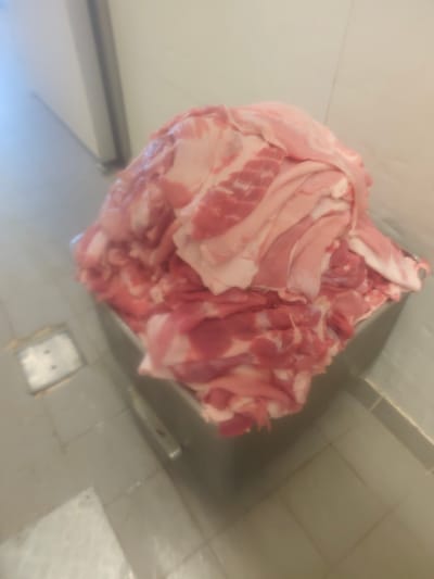 A large pile of raw, thinly sliced pork is contained within a stainless steel bin. The meat is a mixture of pink lean muscle and white fat, layered and folded upon itself. The bin is situated on a tiled floor with visible grout lines. In the background, a light-colored wall and a vertical structure are present. The lighting appears to be overhead and diffused, casting a soft glow on the scene. The image is slightly out of focus, giving it a soft, slightly blurred appearance. The location is identified as the city of Menemeni Municipal Unit, Greece.