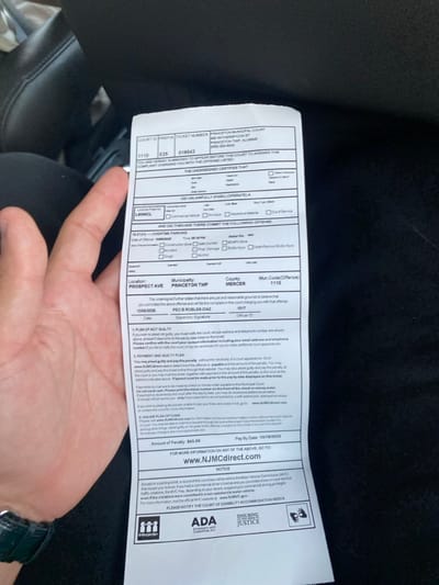 The image depicts a white paper parking ticket held vertically in a person's left hand, visible from the wrist up, inside a vehicle. The background consists of out-of-focus, dark, textured upholstery, likely a car seat.
The ticket is issued by Princeton Municipal Court, located at 400 Witherspoon Street, Princeton, New Jersey, United States. The offense listed is "OVERTIME PARKING" (19:21(P)) which occurred on Prospect Avenue in Princeton Township, Mercer County, on October 6, 2005. The ticket specifies a license plate of L80WCL and an officer identified as PEO B ROBLES-DIAZ (ID: 1017). The penalty amount is $45.00, with a pay-by date of October 19, 2025. The ticket includes instructions for payment via NJMCDirect.com and displays logos for Interpreter, ADA, and Ensuring Justice.