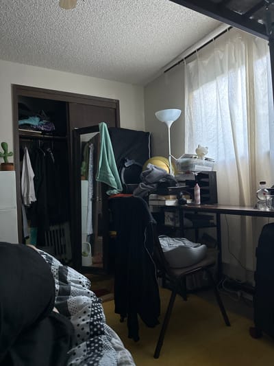 The image depicts a cluttered bedroom. On the left, a closet is partially open, revealing clothes hanging on a rack and items stacked on shelves. A small potted cactus is visible on top of a white dresser next to the closet. In the center of the room, a desk is covered with various items including papers, a lamp, headphones, and bottles. A chair sits in front of the desk, with a gray cushion on it. A large window with sheer curtains is on the right side of the room. The floor is covered with a yellow carpet. A person is partially visible in the foreground, lying on a bed with a patterned blanket. The overall impression is one of disarray and a lived-in space. There is no information in the image to suggest the location is in Vancouver, United States of America.