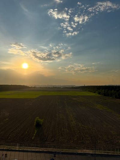 The image captures an aerial view of agricultural fields and a distant city skyline at sunset in Kraskovo, Russia. The sun, a bright orb of yellow, hangs low in the hazy, light blue sky, casting a warm glow over the scene. Wisps of white clouds are scattered across the sky. In the foreground, a vast, dark brown field, recently plowed and showing distinct furrows, dominates the lower half of the image. A lone, small green tree stands out in the middle of this field. Beyond this, a strip of vibrant green agricultural land is visible, likely a growing crop. Further in the distance, a dense line of dark green trees forms a natural boundary. Beyond the treeline, a city skyline stretches across the horizon, marked by numerous buildings and what appear to be cranes, suggesting ongoing construction. The light from the setting sun creates a soft, diffused light across the entire landscape. A dark, paved area with faint lines, possibly a road or parking lot, with a metal fence and some lampposts, is visible at the very bottom of the frame.