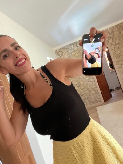 In a room with stone walls, a woman is taking a selfie with her phone. She is wearing a black tank top and a yellow skirt. The phone screen shows a reflection of the woman taking the picture. The room appears to be a bedroom or living area, with a wooden door and a rug on the floor. The location is a private residence in Caldas da Rainha, Portugal.