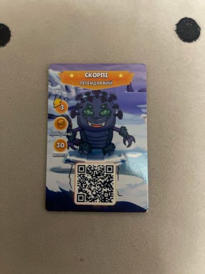 The image shows a collectible card featuring a cartoon monster character. The card has a purple and orange border and depicts a snowy landscape with a purple, multi-eyed monster in the center. Text on the card is in Cyrillic script. There is a QR code at the bottom of the card. The card is lying on a textured, light brown surface, which appears to be a fabric or a similar material. Two dark circular objects are visible on the surface to the left and right of the card. The image is taken from a slightly overhead angle. This scene is not located in the city of Berezhany, Ukraine.