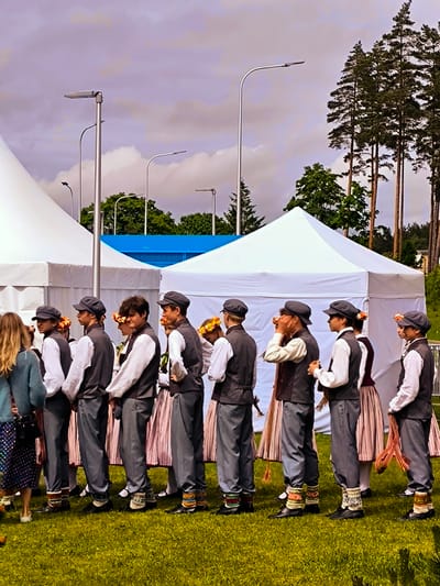 This image captures an outdoor scene in Ulbroka, Latvia, featuring a group of approximately fifteen individuals, primarily young males, standing in two parallel lines on a vibrant green lawn. The males are dressed in dark grey vests over white long-sleeved shirts, dark grey trousers, and dark peaked caps; many exhibit brightly patterned, knitted ankle socks above their black shoes. Interspersed within the group are several females wearing long, striped or patterned skirts, and some adorn floral wreaths on their heads. On the far left, a blonde-haired female, seen from behind, wears a blue-green long-sleeved top and a multi-colored patterned skirt.
In the background, multiple white canvas event tents of varying sizes are erected. A large white tent occupies the left side, with additional smaller white tents extending towards the right. A distinct blue structure is visible between two tents in the mid-background. Several modern, grey streetlights with curved poles extend upwards across the upper frame, positioned above the tents. Tall pine trees are visible on the right side and in the distant background, alongside other deciduous foliage. The sky above is overcast with a mix of grey and purple-tinged clouds. The individuals appear to be in a waiting posture, uniformly oriented generally towards the right, suggesting a pre-event assembly or a ceremonial lineup.
