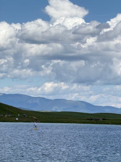 The image depicts a wide outdoor scene in Javorje, Montenegro, featuring a lake, green hillsides, distant mountains, and an overcast sky. The sky is dominated by large, white and grey cumulus clouds, with intermittent patches of blue visible primarily in the upper sections. A faint contrail is present in the upper right quadrant of the sky. In the background, a range of hazy blue-grey mountains stretches across the horizon. The midground consists of gently sloping green hills, covered in grass. On the left hillside, several indistinct dark shapes, possibly livestock or rocks, are scattered. A dark-colored vehicle is parked on this hillside, near at least two individuals, one in a white garment and another darker-clad figure. On the right midground, closer to the lake, a small, dark, dilapidated structure, possibly an old shed or dock, is visible. The foreground is occupied by a large body of water, likely a lake, exhibiting a rippled, dark blue-grey surface. In the center of the lake, a single individual, wearing a white top and light-colored shorts, stands on a bright yellow stand-up paddleboard (SUP) and holds a paddle. The person appears to be actively paddling or maintaining balance on the board. Further to the right on the shore, another individual is seen bending over near the water's edge.