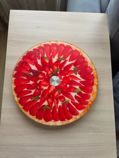 A circular strawberry tart, depicted in a high-quality photograph, is centrally placed on a light-colored wooden table. The tart is adorned with numerous sliced strawberries, arranged in a radial pattern, with some mint leaves interspersed among them. The crust of the tart is visible at the edge, appearing golden brown. In the center of the tart, a silver metallic element with a spiral pattern is present. The table surface exhibits a subtle wood grain texture. To the right of the table, a portion of a grey upholstered chair is visible, and further to the right, dark fabric, possibly denim, can be seen. The background includes a blurred view of vertical blinds. The overall scene is well-lit, suggesting an indoor setting. The image does not contain any identifiable landmarks or elements that would place it specifically in Скалба Снт, Russia.