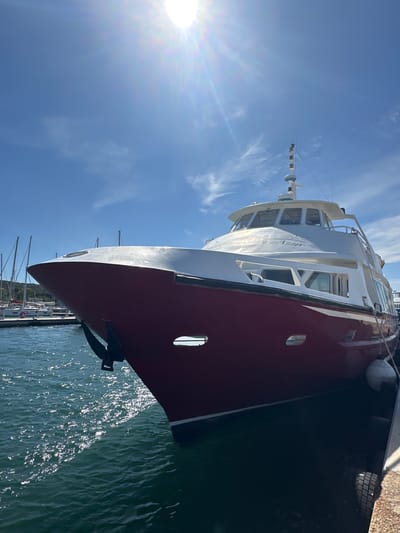 A large, red and white ferryboat named "Belle de Jour" is docked in the port of Hyères, France. The boat is positioned at an angle, with its bow facing towards the viewer and to the left. The sun is shining brightly, casting a lens flare across the blue sky. The water is calm and reflects the sunlight. In the background, other sailboats and yachts are visible, as are the green hills surrounding the port. The ferryboat has multiple windows, and a small antenna on its roof. The scene is a typical summer day in a Mediterranean port.