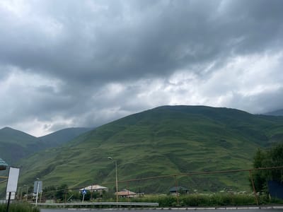 This image captures a landscape in Arsha, Georgia, characterized by rolling green mountains under a heavily clouded sky. The foreground features a road with a white dividing line, bordered by a guardrail and overgrown grass. Several houses, appearing to be rural dwellings with tiled roofs, are visible in the middle ground. A street lamp stands near the road, and various signs are present, including a blue and white pedestrian crossing sign. The dominant feature is the expansive, verdant mountain slopes, etched with subtle contours and shadows, indicative of natural terrain. The overall atmosphere is subdued due to the overcast weather.