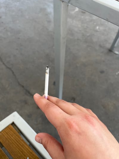 A person's hand holds a lit cigarette with ash at the tip. The cigarette has a white paper filter and a pink tip. The hand is positioned in the foreground, with the fingers visible. In the background, a blurry concrete or asphalt surface is visible, along with the leg of a metal bench. The overall impression is of an outdoor setting, possibly a public space. There are no other people visible in the image. The location in Gorni Dabnik, Bulgaria is not discernible from the image itself.
