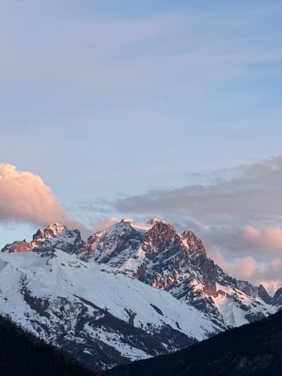 The image shows a mountain range with snow-covered peaks and rocky outcrops, illuminated by the setting sun. The sky is pale blue, with some pinkish clouds near the horizon. The location is L'Argentière-la-Bessée, France. The mountain range is the prominent element, its snow reflecting the sunlight and creating a contrast with the dark rocky areas. The image is rotated.