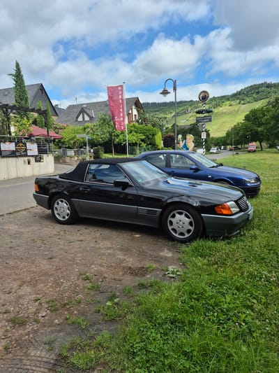 This image captures a daytime scene in Traben-Trarbach, Germany, specifically in the vicinity of the "Moselperle" restaurant on Gartenstrasse.

In the foreground, a black Mercedes-Benz R129 SL-Class convertible with a raised black soft top is parked on a dirt and gravel patch, partially overlapping with a grass verge on its right side. The vehicle features grey lower body cladding and silver multi-spoke wheels. Behind and slightly to the right of the Mercedes, a dark blue sedan, appearing to be a Seat Toledo 1M, is parked, primarily situated on the grassy area. The ground beneath the vehicles consists of loose earth, gravel, and sporadic patches of green grass, displaying tire tracks in the dirt.

The mid-ground features several buildings with dark grey pitched roofs, some equipped with dormer windows, suggesting residential or hospitality structures. A red vertical banner, partially visible, reads "MOSELPERLE WEINRESTAURANT." To the left, a sign on a building also indicates "MOSELPERLE." A lamppost stands near a signpost displaying "Gartenterrasse Moselperle" below a circular emblem, and further down, a cartoon-like figure holding a glass. A blue street sign, "Gartenstrasse," is visible. Lush green foliage, including bushes and trees, surrounds the buildings and lines the paved road to the left of the parked cars.

The background reveals a steep hillside characteristic of the Mosel Valley, extensively cultivated with vineyards and topped with dense forest. The sky above is partly cloudy, featuring white and grey clouds interspersed with areas of clear blue. In the far distance, on a road at the base of the hillside, a white car and a red van are visible. No people are present or engaged in discernible activities within the frame.