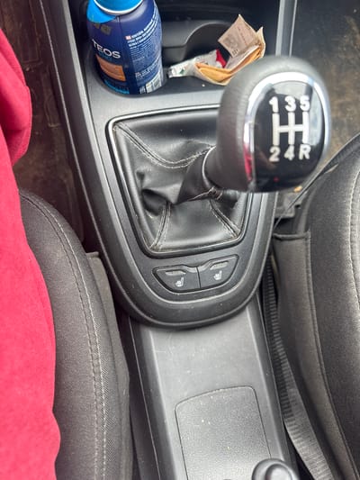 The image shows the interior of a car, focusing on the gear shifter and the center console. A red piece of fabric, likely clothing, is visible on the left side of the image, draped over a seat. The gear shifter has markings for gears 1, 3, 5, 2, 4, and R. Below the gear shifter, there are two buttons with icons resembling heated seats. To the right of the gear shifter, a cup holder contains a blue bottle with the brand name "TEOS PRO" visible, and a crumpled piece of paper. The overall environment appears to be a car's interior, possibly parked or in motion. There are no visible people in the image. The location is specified as the city of Chastyye, Russia.