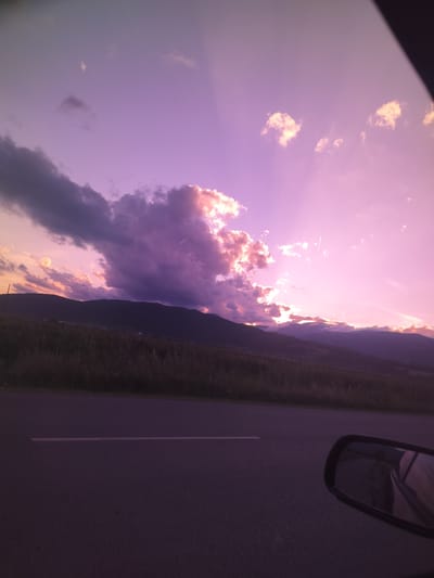 The image depicts a sunset over a rural landscape, viewed from the perspective of a vehicle. The sky is a vibrant gradient of pink and purple, with distinct sunbeams emanating from behind the clouds. A large, dark cloud dominates the left side of the sky, its edges illuminated by the setting sun. Below the sky, a range of dark, silhouetted mountains stretches across the horizon. In the foreground, a paved road with a white line is visible, with a portion of a car's side mirror reflecting the scene on the right side of the frame. A strip of vegetation, appearing dry and possibly overgrown with grass and wildflowers, lies between the road and the mountains. The overall impression is one of a serene, natural landscape at dusk, likely in the vicinity of Manolovo, Bulgaria, given the provided context.
