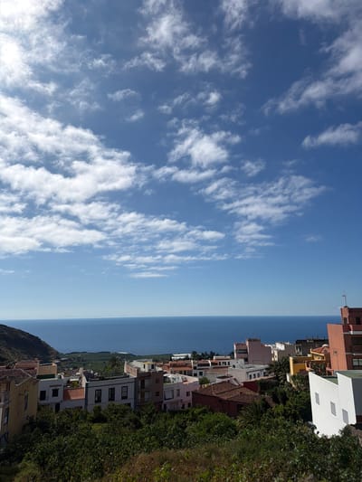 This is a photograph taken from an elevated viewpoint overlooking the coastal town of Icod De Los Vinos, Spain. The image is dominated by a clear blue sky with scattered white clouds, a common sight in the Canary Islands. Below the sky, the vast expanse of the deep blue Atlantic Ocean stretches to the horizon.
In the midground, a cluster of buildings forms the town's skyline. The architecture is varied, featuring multi-story structures with flat roofs and some with traditional terracotta tiled roofs. The buildings are painted in a range of colors, including white, cream, pale pink, and a prominent salmon pink. Some buildings have visible windows and balconies, suggesting residential or commercial use. The buildings are nestled amongst lush green vegetation, with trees and bushes encroaching on the built environment.
Further down the hillside, the landscape transitions to terraced agricultural fields, likely banana plantations, which are a significant crop in the region. The foreground is filled with dense, dark green foliage, obscuring the very bottom edge of the image. The overall impression is one of a sunny day in a coastal town, with a clear demarcation between the sky, sea, town, and vegetation. There are no people or visible activity captured in the frame.
