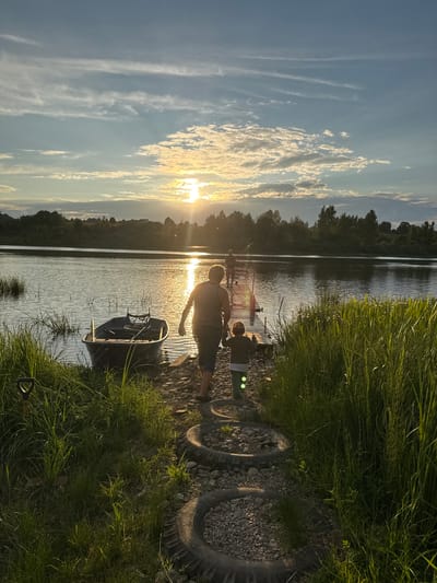 This is a full, outdoor, vertical, eye-level shot of a riverbank scene at dusk. The sun is setting to the left, casting a warm glow on the water and sky. The sky is a mix of blue and white clouds, with streaks of cirrus clouds high up.
In the foreground, tall green grass lines the right side of the frame. A pathway made of old tires filled with gravel leads from the bottom of the image towards a wooden pier. A shovel is stuck in the ground to the left of the pathway.
On the left side of the image, a small, dark-colored boat is moored by the bank. The water is calm with ripples reflecting the sunlight.
On the wooden pier, two people are visible. An adult, silhouetted against the setting sun, walks away from the camera towards the water. They are wearing a striped tank top and dark shorts. A child, also silhouetted, walks slightly ahead of the adult, holding their hand. The child is wearing a green shirt and pants. Further down the pier, another person is visible, possibly fishing, as they are holding a fishing rod.
The background consists of a dense line of trees and foliage along the opposite bank of the river. The overall atmosphere is peaceful and serene, capturing a moment of outdoor activity at sunset. The location is identified as Sitno, Russia.