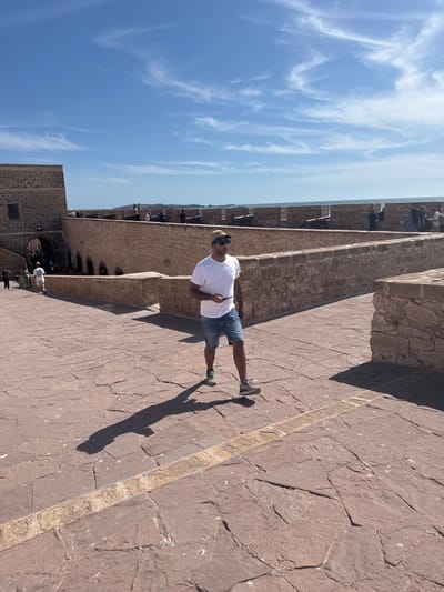 A man in a white t-shirt, denim shorts, sunglasses, and a baseball cap walks across a large, paved courtyard. He is holding a phone in his right hand and casting a long shadow to his left. The courtyard is made of reddish-brown stone tiles. In the background, a stone wall with cannons is visible along the edge of a fortified structure. Beyond the wall, the blue ocean meets the horizon under a bright blue sky with wispy clouds. Other people are scattered in the background, some standing near the wall and others walking near an arched gateway. The scene appears to be within the ramparts of the Skala de la Ville in Essaouira, Morocco, a historic coastal fortification.